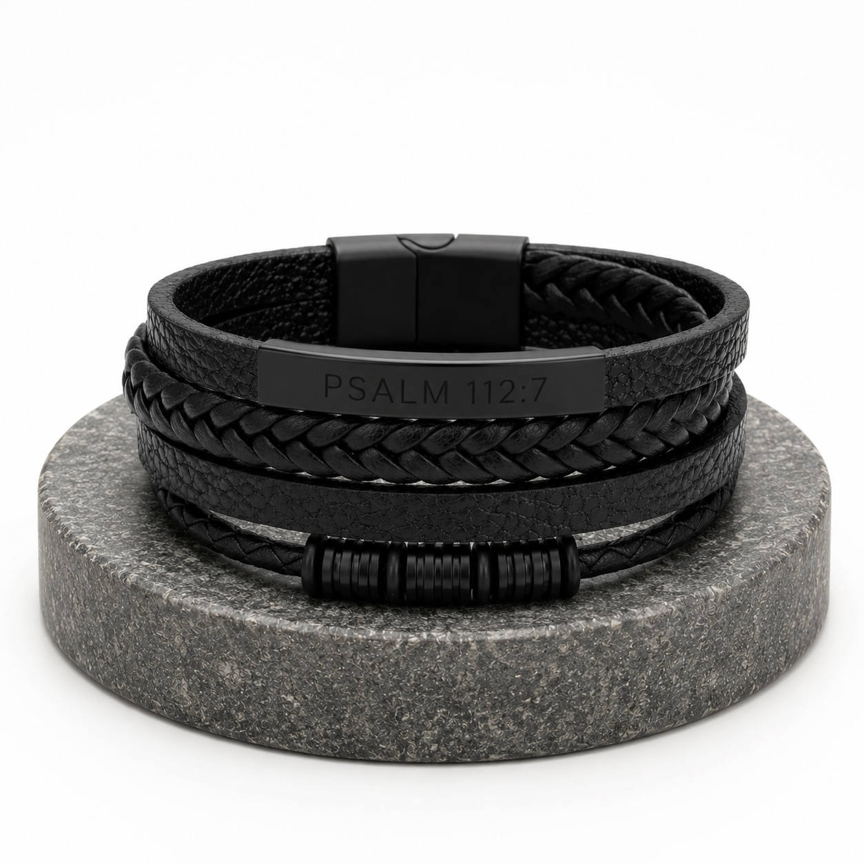 A stack of black leather bracelets, including braided and smooth woven styles, displayed on a round gray stone stand against a white background. One bracelet features a black metal plate engraved with 'PSALM 112:7'.