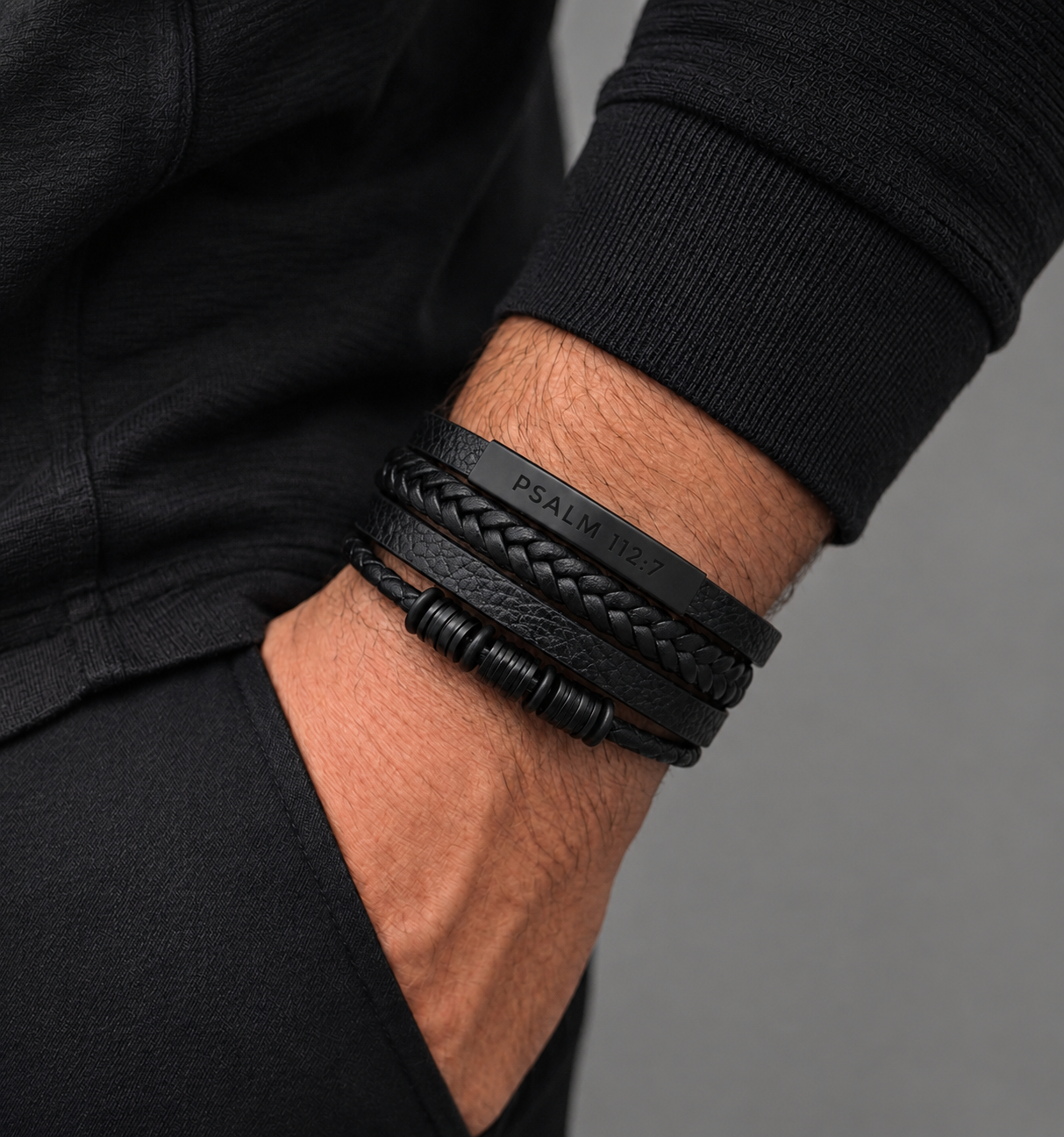 A close-up of a person's wrist wearing multiple black leather bracelets, including braided and smooth bands, against a dark gray background.